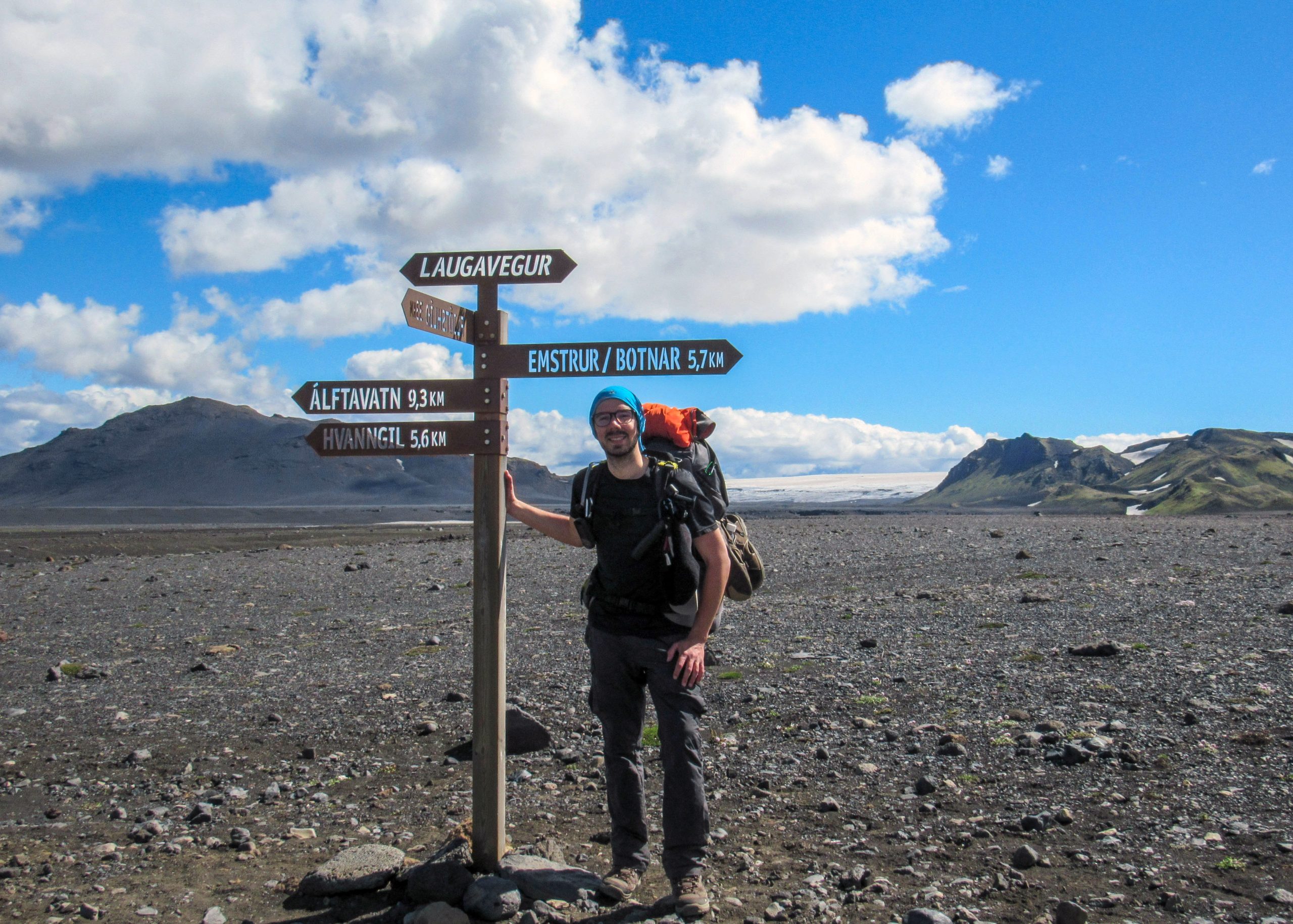 Experienced hiker with heavy backpack staying next to navigation signpost and enjoying beautiful scenery of volcanic mountains and glaciers, Iceland